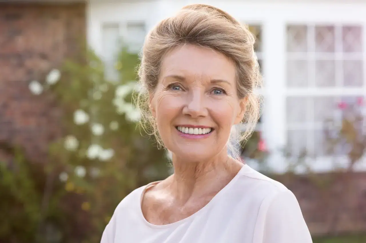 Visage femme 80 ans souriant, cheveux blonds relev&eacute;s, devant une maison.