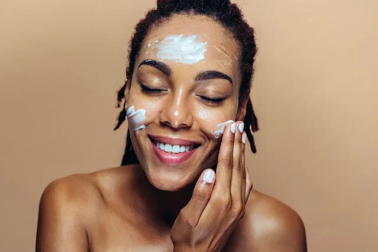 Une femme souriante se met de la crème sur le visage, les yeux fermés, pour un soin de peau doux et hydratant.