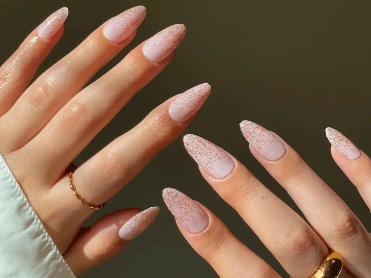 Mains délicates aux ongles paillettes dégradé rose pâle, ornées de bagues fines.