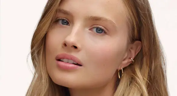 Visage d'une jeune femme aux yeux bleus et aux lèvres rosées, avec un maquillage subtil, style "makeup no makeup". Elle porte des boucles d'oreilles dorées.